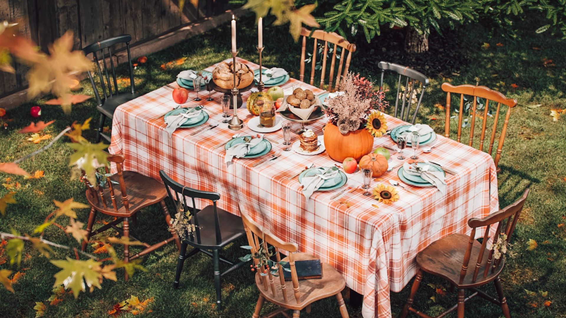Harvest-themed outdoor dining experience replete with the enchantment of fall, featuring pumpkin centerpieces and a checkered orange tablecloth amid a flurry of fallen leaves.