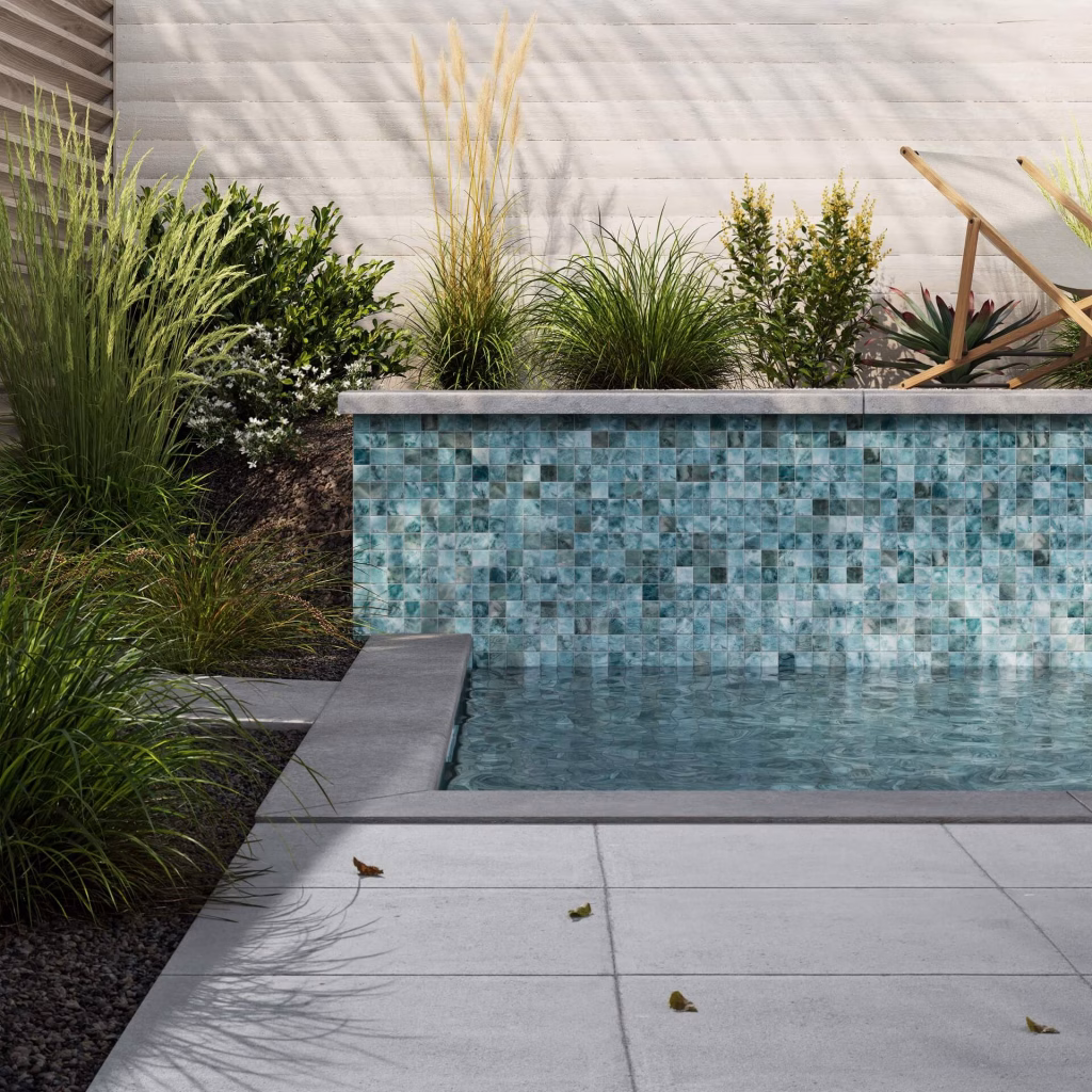 3D render of a garden wall of teal glass mosaic squares rising from a small plunge pool, with ornamental grasses and a deck chair